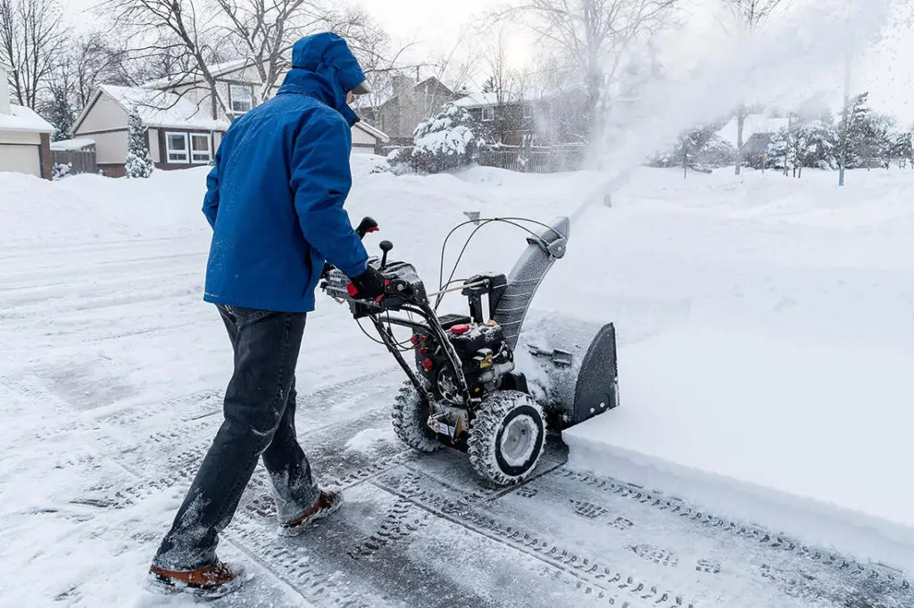 A worker using a snow blower removes snow from a residential lot.